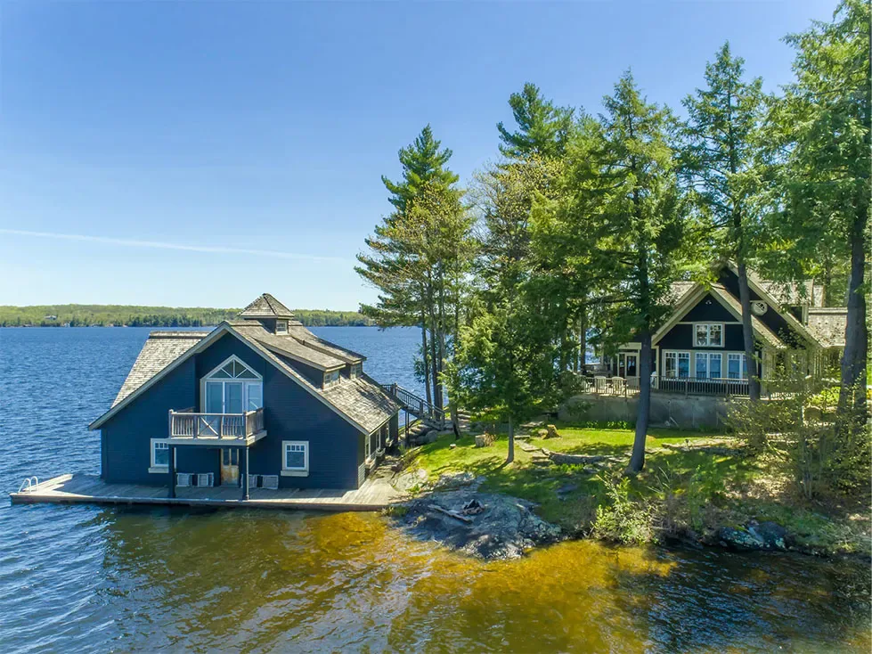 A blue boathouse on a rocky point, with shallow lake water in front and the bottom visible through it
